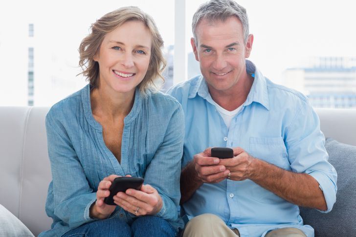 Happy couple using their smartphones looking at camera at home i