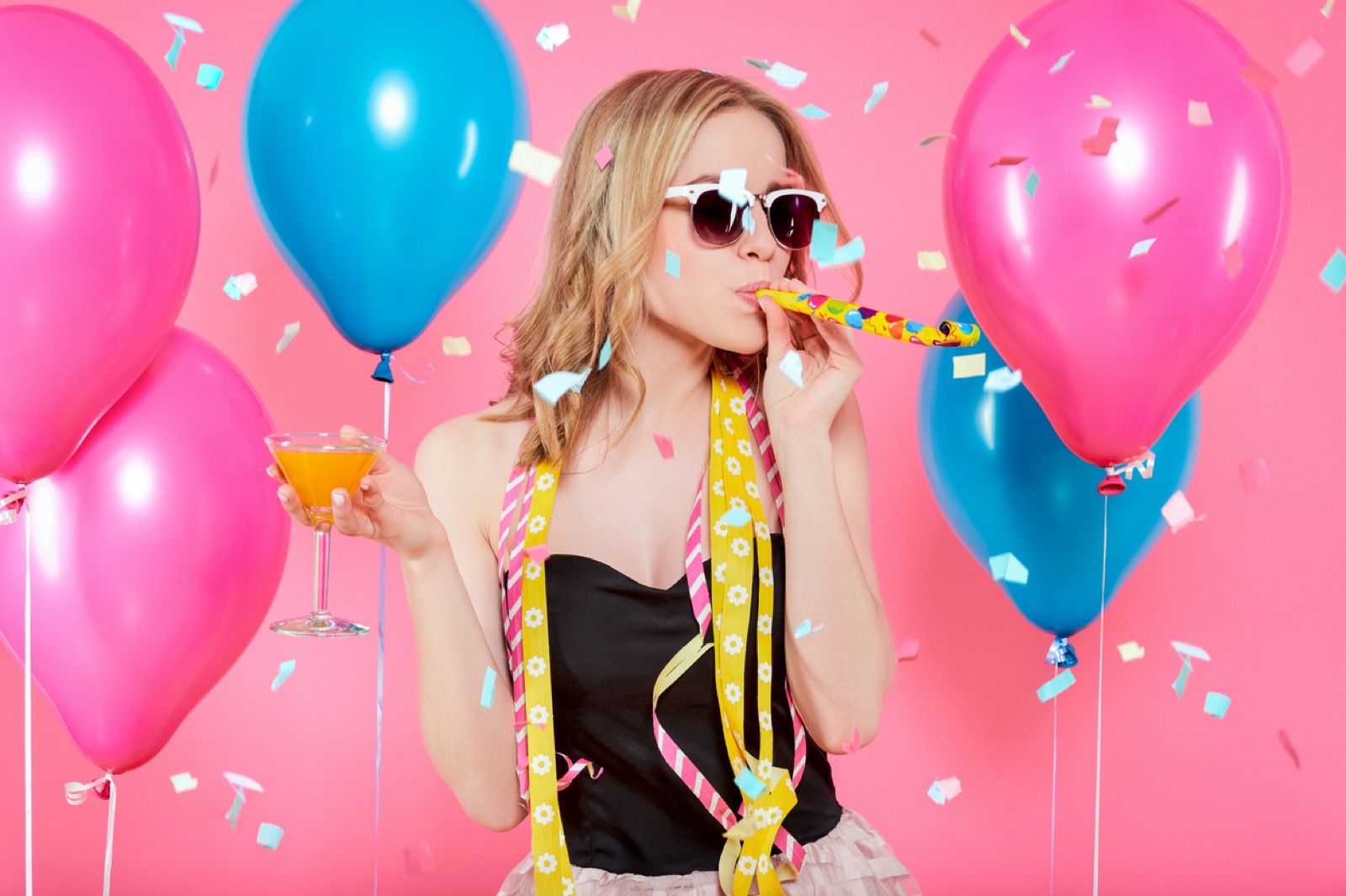 😠 What % Antisocial Are You? Gorgeous Trendy Young Woman In Party Outfit Celebrating Birthday. Party Mood, Balloons, Noisemaker, Flying Confetti, Cocktail And Dancing Concept On Pastel Pink Background.