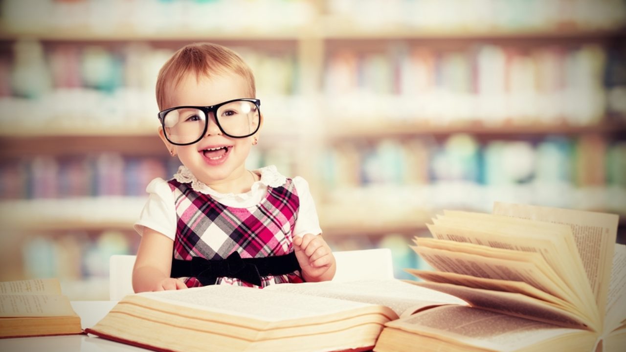 Is Your Vocabulary Better Than the Average Person? Smart Baby Girl In Glasses Reading A Book In A Library