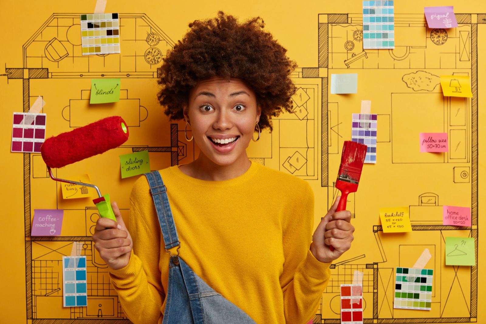 Creative Painter Holds Paint Roller And Brush, Does House Repairing, Dressed In Yellow Jumper And Overalls, Stands Against House Sketch With Color Samples, Sticky Notes. Renovation